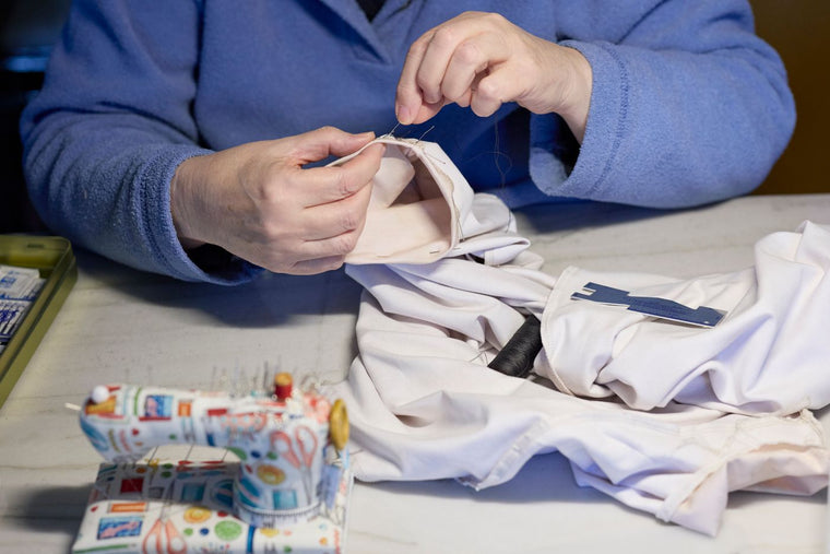 Person in a blue jacket hand-stitching a white garment beside sewing tools